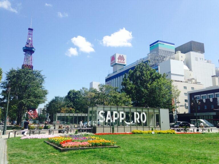 Odori Park in Sapporo, Japan, with a sign saying "Sapporo". The Iconic Sapporo TV Tower is seen in the distance. The park has green grass along with blooming flowers and trees.