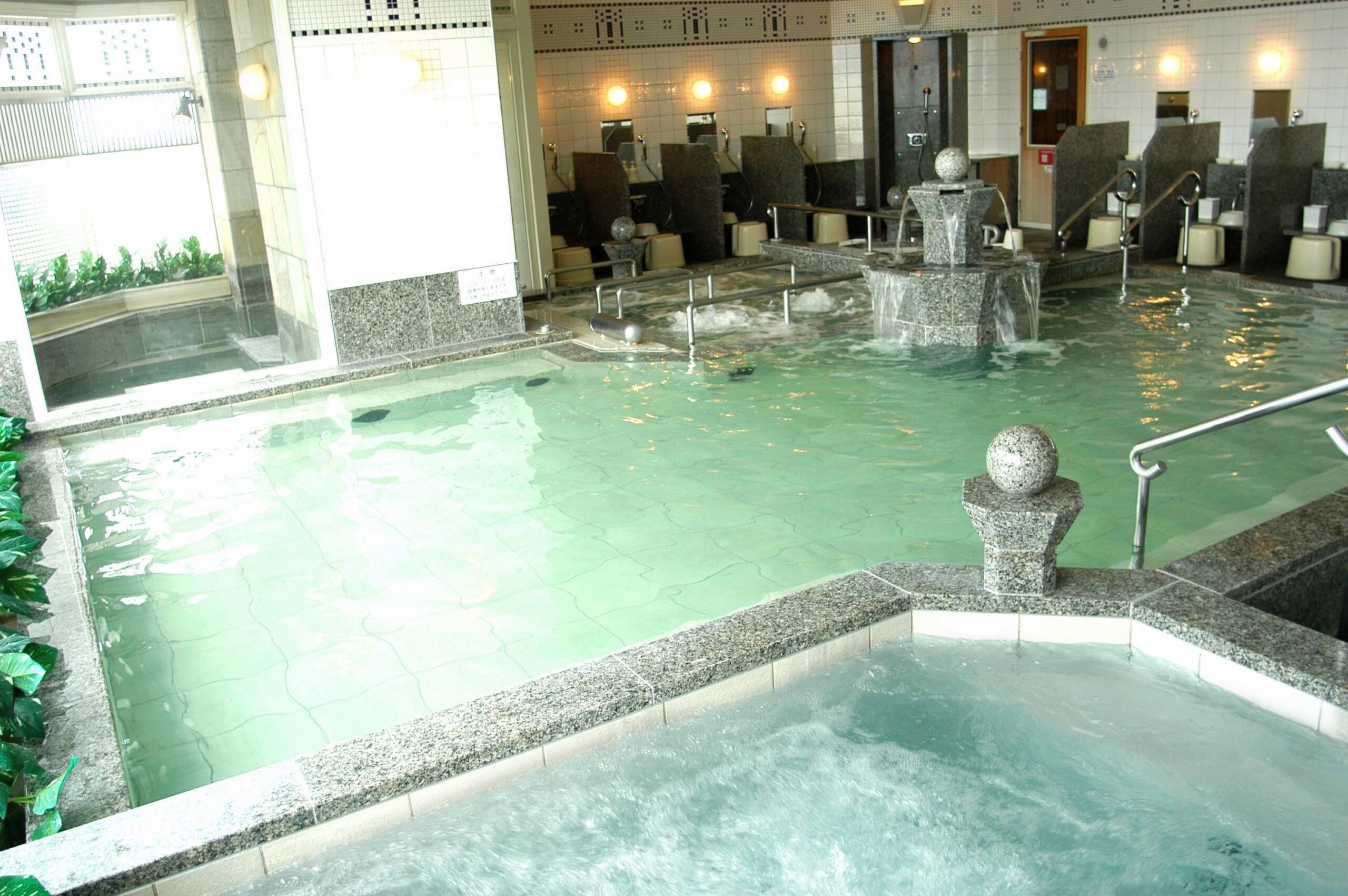 A large public onsen with clear water and a fountain in the pool. The onsen is available at Hotel Monterey Edelhof in Sapporo, Japan.