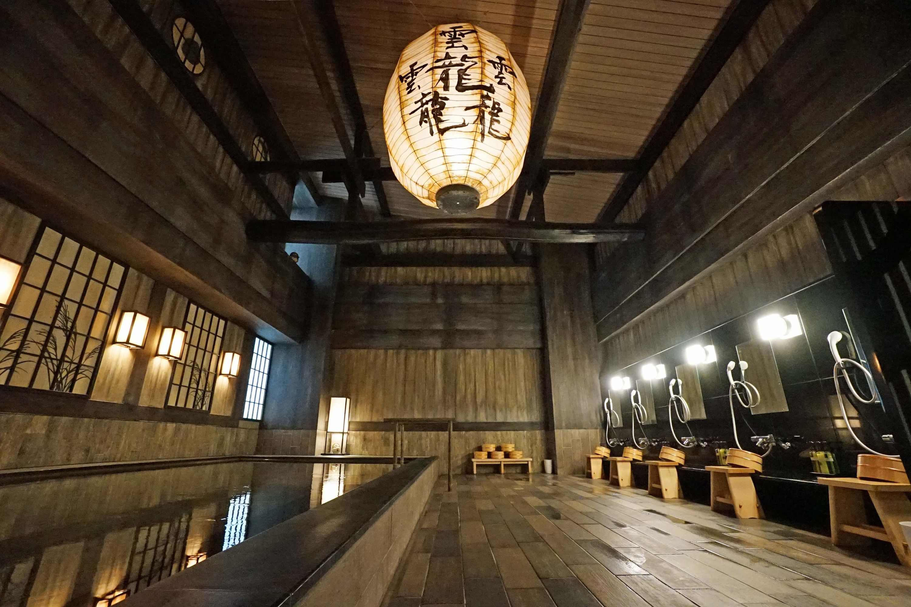 A Japanese-style onsen at La'gent Stay Sapporo, a hotel in Japan. There is a large onsen to the left, with public showers to the right. There is a large lantern illuminating the room in the middle.