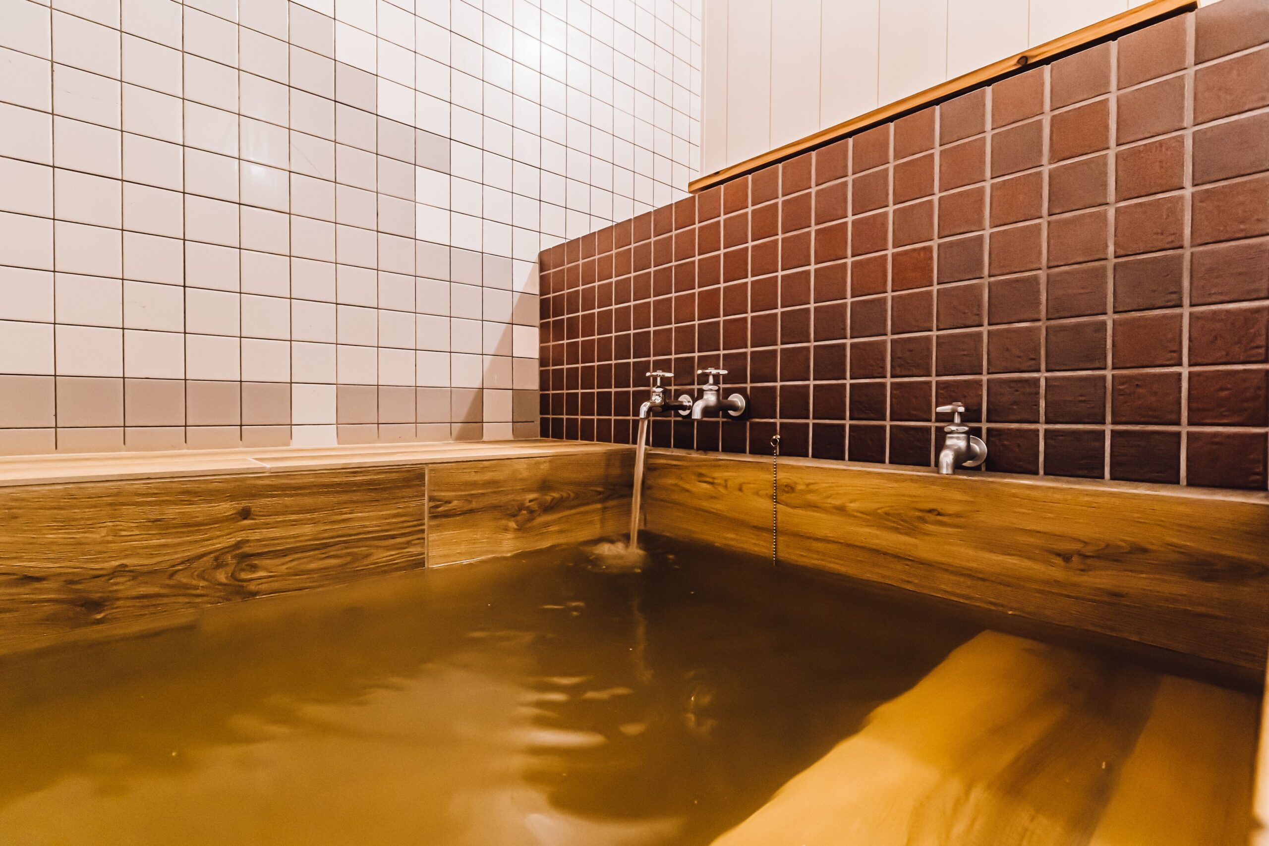 A large onsen made of wood with a faucet pouring hot spring water. There are brown tiles and white tiles decorating the bathroom. This onsen is available at Yamahana Onsen Tonden Japanese Inn.