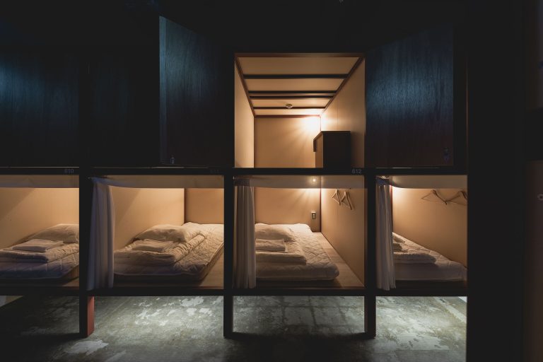 A deluxe capsule pod in Helio Hostel, located in Sapporo, Japan. Four beds are shown, and a door is open showing the ample overhead space ideal for storing luggage.