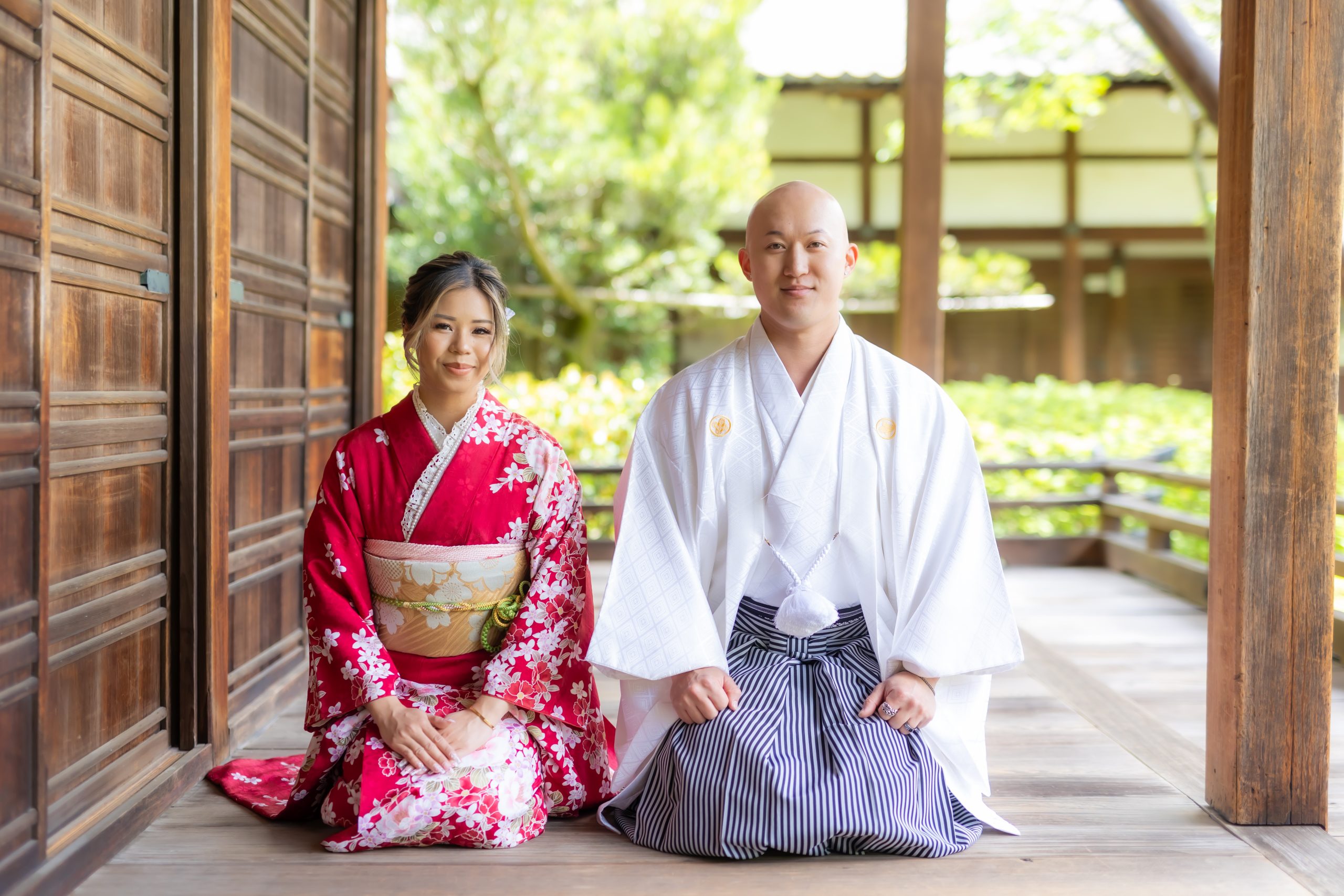 A couple dressed in traditional Japanese attire sits on a wooden veranda. The woman is wearing a vibrant red kimono with floral patterns, while the man is dressed in a white ceremonial kimono with a striped hakama. They are both sitting in a serene, traditional setting with a lush garden in the background, creating a calm and elegant atmosphere.