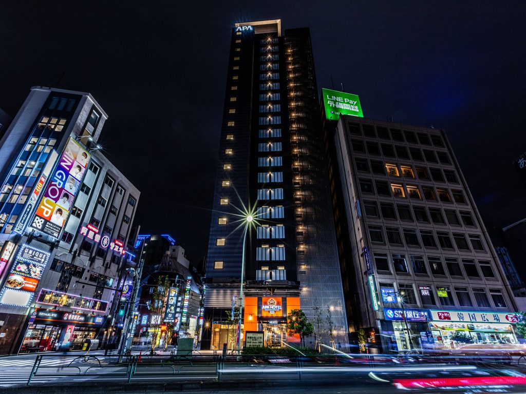 A street view of APA Hotel Higashi Shinjuku Kabukicho Tower in Tokyo, Japan at night. It is a high rise hotel with many floors.
