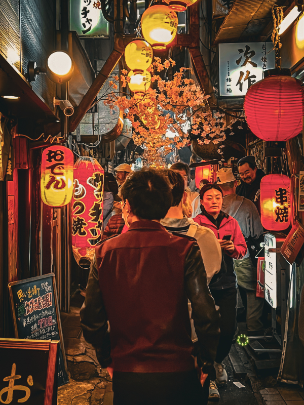 A narrow, bustling alleyway filled with people walking through a vibrant and cozy night scene in Tokyo, Japan. The area is illuminated by numerous colorful paper lanterns with Japanese characters, hanging from the buildings on both sides. Cherry blossom decorations add a warm touch to the ambiance. People are dressed in casual clothing, with the crowd giving the scene a lively, inviting feel. The signs, lanterns, and warm lighting create an authentic Japanese street market atmosphere in Nonbei Yokocho.