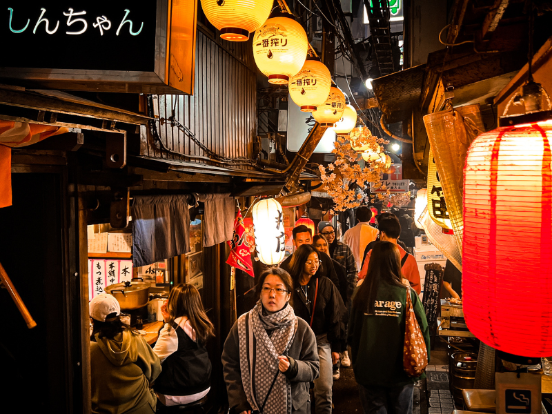 A narrow, bustling alleyway filled with people walking through a vibrant and cozy night scene in Tokyo, Japan. The area is illuminated by numerous colorful paper lanterns with Japanese characters, hanging from the buildings on both sides. Cherry blossom decorations add a warm touch to the ambiance. People are dressed in casual clothing, with the crowd giving the scene a lively, inviting feel. The signs, lanterns, and warm lighting create an authentic Japanese street market atmosphere in Nonbei Yokocho.
