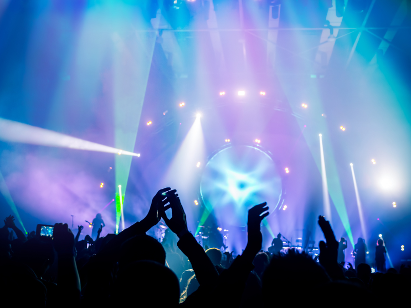 A vibrant nightlife scene in Tokyo, with a crowd enjoying live music under colorful stage lights. The silhouettes of people raising their hands and dancing create an energetic atmosphere, while neon blue and purple lights illuminate the venue, adding to the electric vibe. This image captures the lively spirit of Tokyo’s nightlife.