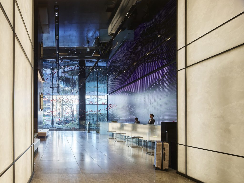 A modern, spacious lobby at Millennium Mitsui Garden Hotel, featuring high ceilings and a sleek, minimalist design. The illuminated reception desk has two staff members ready to assist, while a large digital art display on one wall adds a stylish touch to the space. Natural light streams through the glass walls, offering a view of the outside. A suitcase by the reception area creates a welcoming atmosphere for guests arriving at the hotel.
