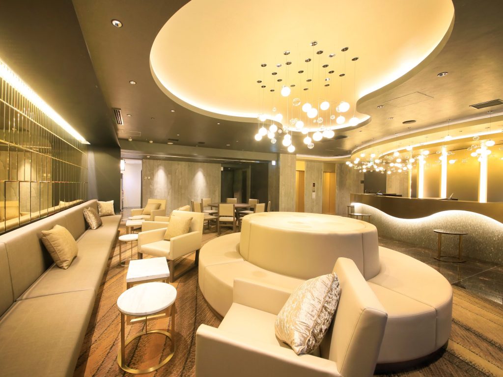 The elegant lobby of Quintessa Hotel Tokyo Ginza featuring warm, ambient lighting, a modern circular seating area, sleek marble flooring, and contemporary furniture with gold accents. The reception desk is illuminated with stylish pendant lights, creating a sophisticated atmosphere.