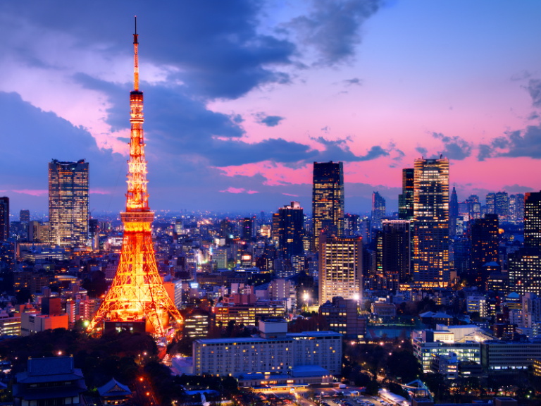 Tokyo Tower illuminated against the evening sky in Japan, with the city skyline stretching out beneath a vibrant sunset of pink and blue hues. The tower's warm lights stand out amidst modern skyscrapers and buildings, creating a stunning contrast that highlights Tokyo's blend of traditional and contemporary charm.