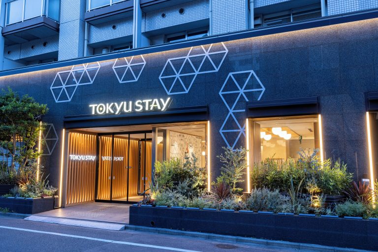 The exterior of Tokyu Stay Tsukiji, a 3 star hotel right next to Tsukiji Outer Market in Tokyo, Japan. The sign says Tokyu Stay, with black paint. Inside is a well lit, modern interior with natural wood and plants around.