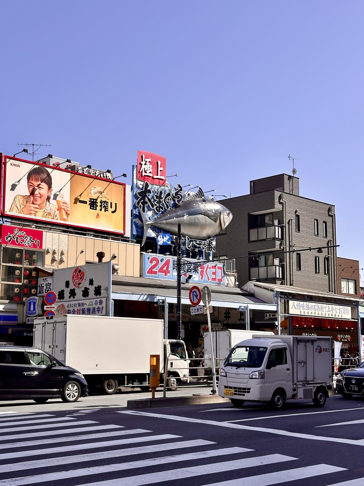 Entrance to Tsukiji Market on a sunny day, featuring the iconic fish billboard sign. The market is visible across a crosswalk, blending subtly into the bustling surroundings.