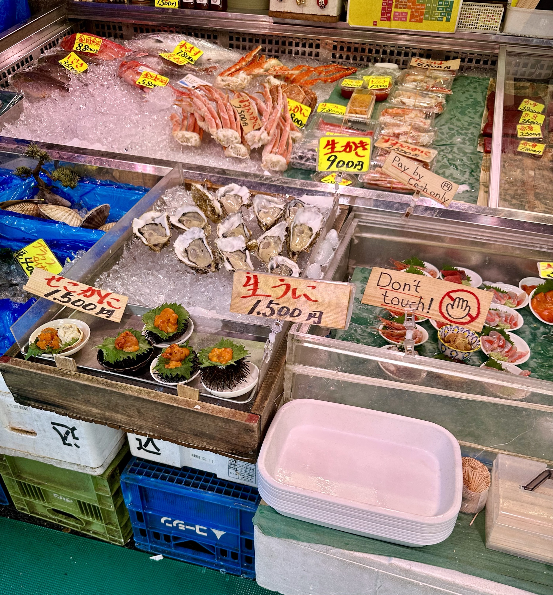 A food vendor stall at Tsukiji Market displaying crab legs, fresh sashimi, and uni. Signs reading 'Cash Only' and 'Don't Touch' are prominently placed above the vibrant seafood selection.