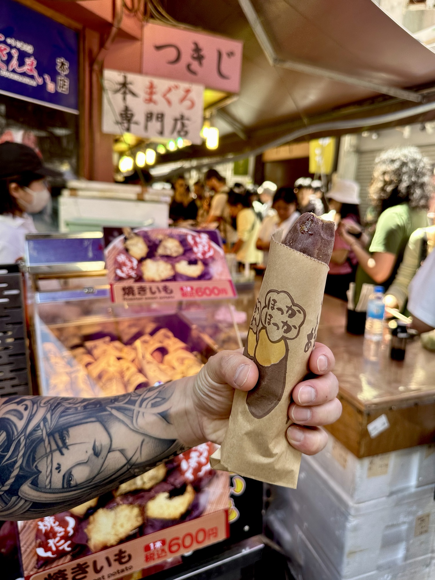 A hot steaming sweet potato from a food vendor in Tsukiji Market. Delicious snacks are being offered at a stall in the background.