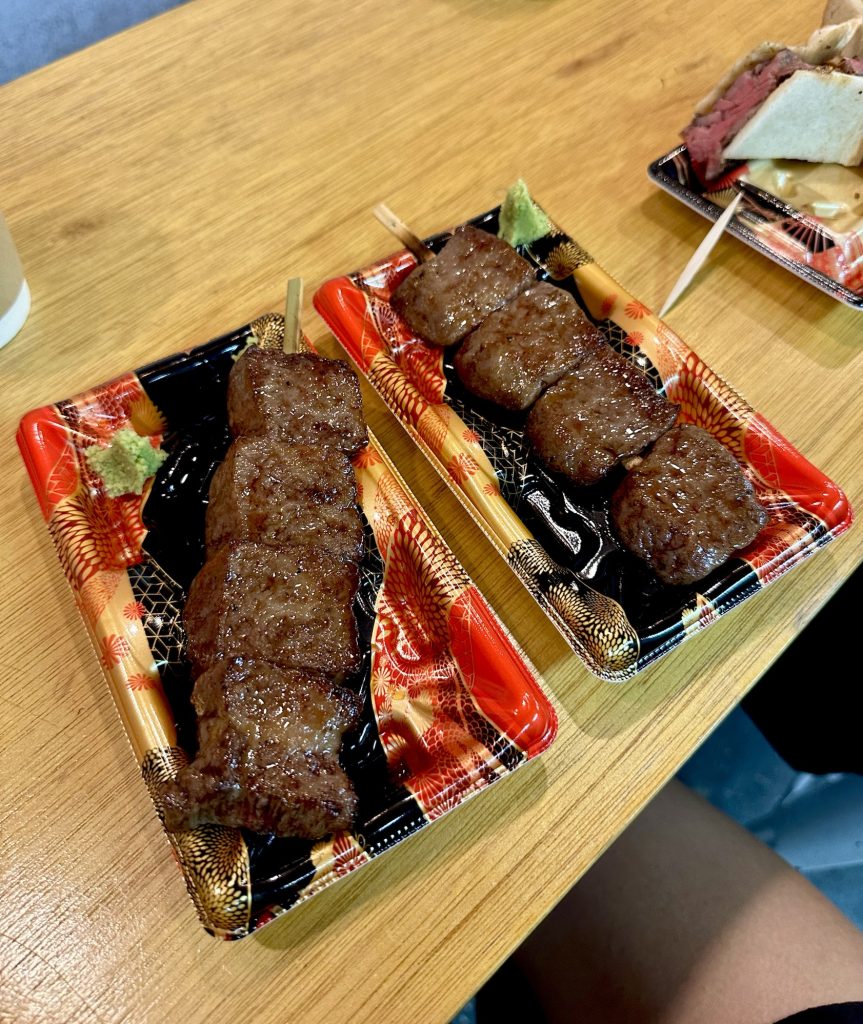 Two perfectly grilled wagyu beef skewers, glistening with juices, served hot from a food vendor stall in Tsukiji Market.