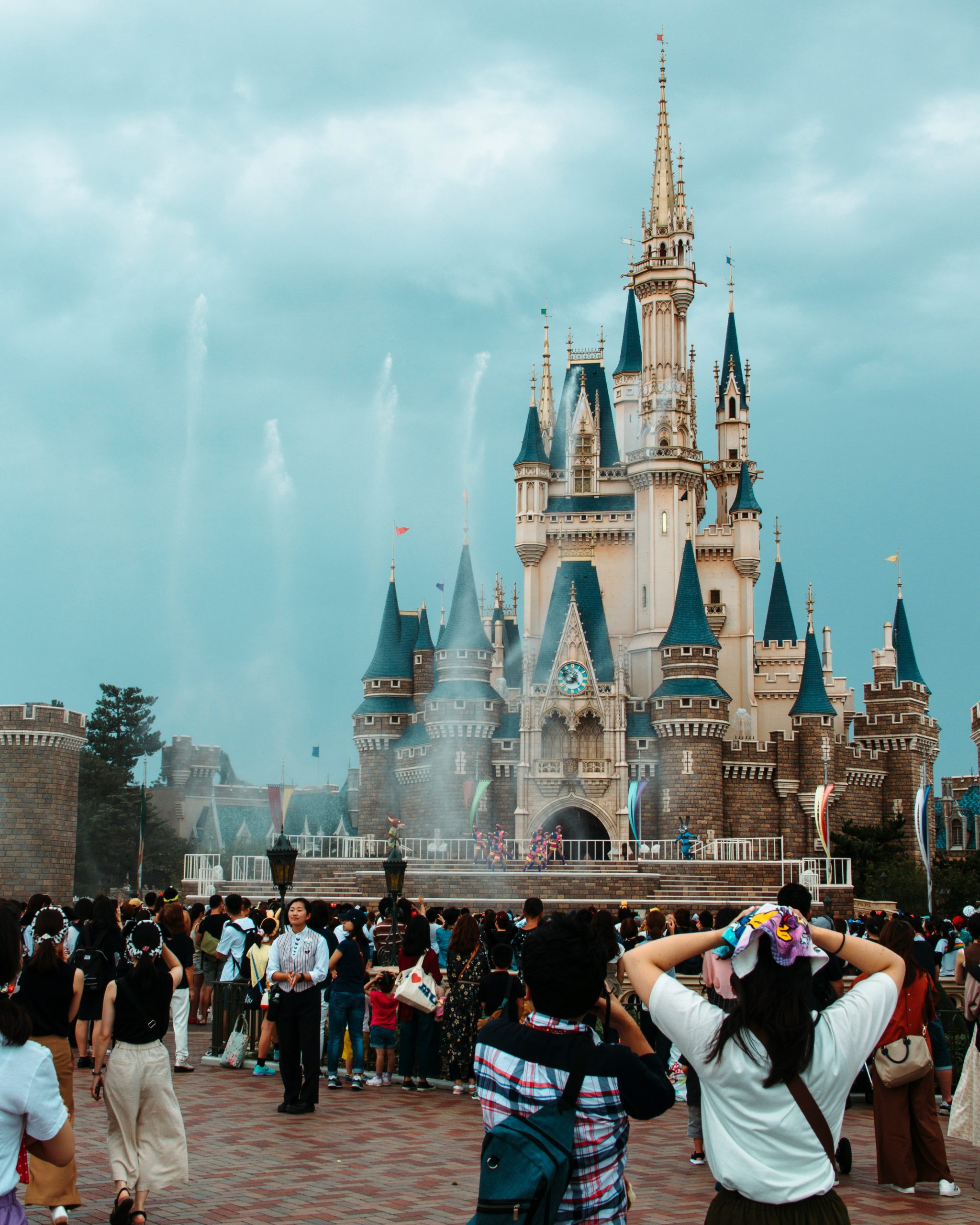 A crowd gathers in front of Cinderella Castle at Tokyo Disneyland, with people excitedly taking photos and enjoying the magical atmosphere. The iconic castle stands tall with its blue and gold spires, set against a cloudy sky, as fountains and decorations add to the enchantment of the scene in this popular theme park.