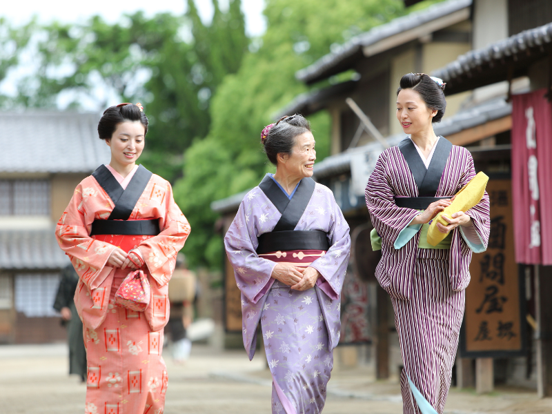 Three women dressed in traditional kimonos walk together, smiling and enjoying the historic setting of Edo Wonderland in Japan. The backdrop features classic Japanese architecture, transporting visitors back to the Edo period and adding to the cultural authenticity of this immersive theme park experience.