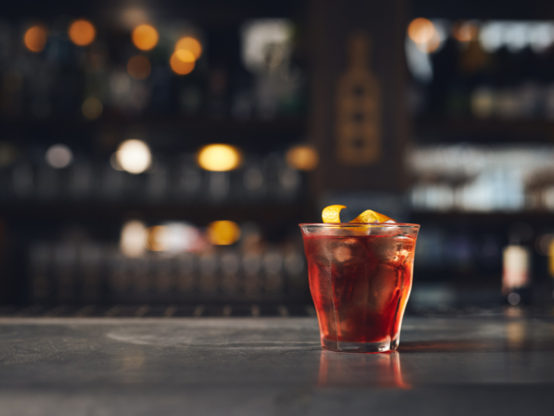 Red cocktail in a glass with a decorative garnish, set against a blurry city skyline in the background.