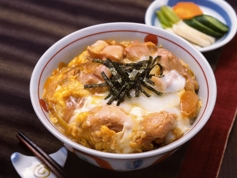Close-up of a traditional oyakodon bowl, showcasing the creamy integration of chicken and egg, crowned with strips of nori seaweed and a side of colorful pickled vegetables - a quintessential Tsukiji Market breakfast.