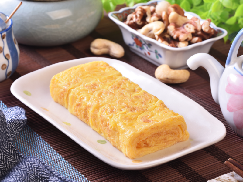 A fluffy roll of golden tamagoyaki served on a white plate, its soft layers glistening. A light and savory breakfast option popular at Tsukiji Market.