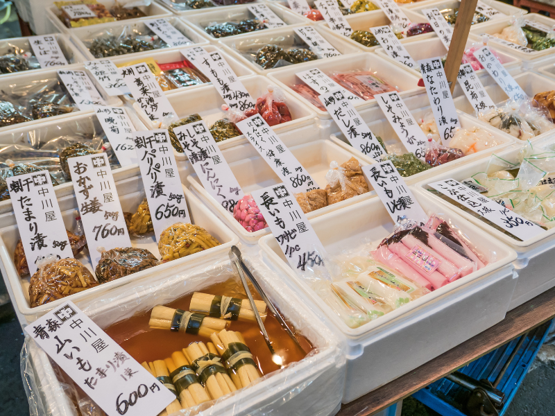 Close-up of assorted foods in white containers at Tsukiji Market, showing different shades of pink, yellow, and brown, with Japanese handwritten price labels visible nearby.