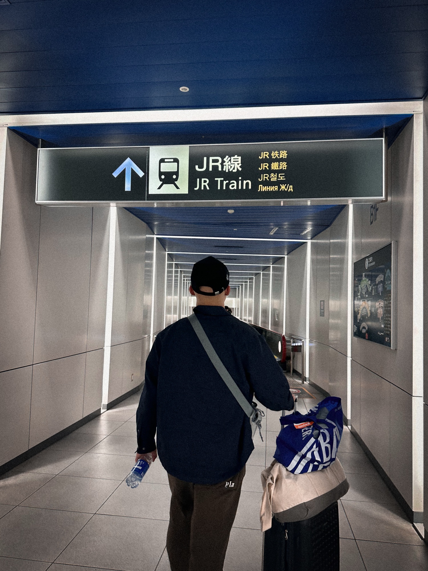 A man with a suitcase walking to the JR Train in New Chitose Airport. There is a sign with a train symbol, saying JR.