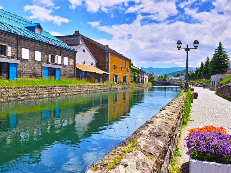 An outdoor view of the Otaru Canal in Otaru, a port town that is a popular day trip from Sapporo. The canal is a bright blue color, with brightly colored houses on the other side.