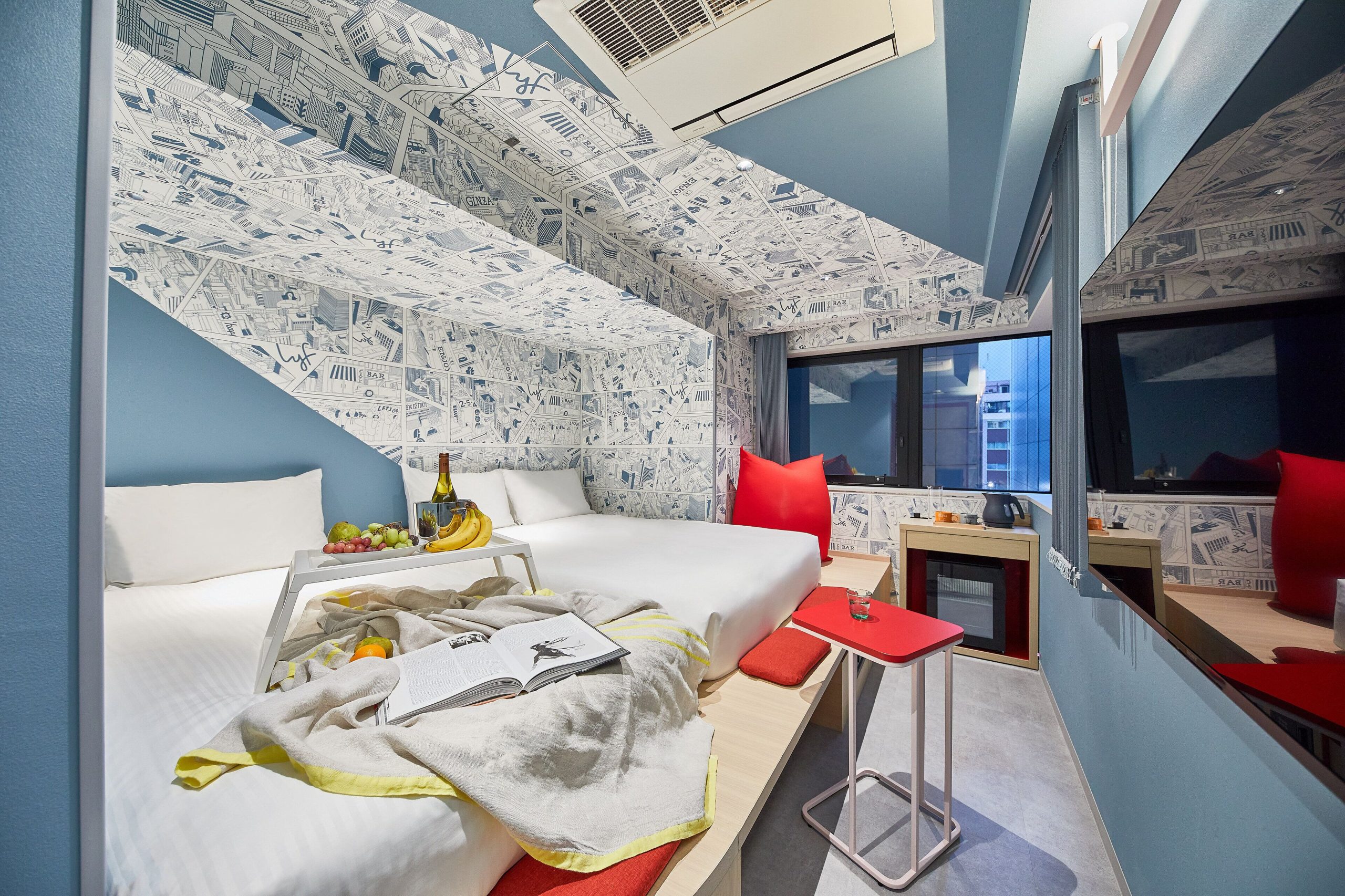 Modern hotel room at lyf Ginza Tokyo featuring two beds, vibrant red accents, comic-style wallpaper, and a city view through large windows.