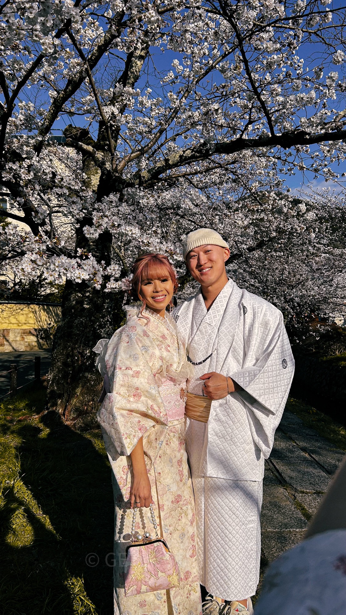 A man and woman wearing kimonos at the Philosopher's Path in Kyoto, Japan. They are smiling at the camera. The girl is wearing a light pink kimono that matches her pink hair, while the man wears a white kimono. Cherry blossoms surround the couple in the background.