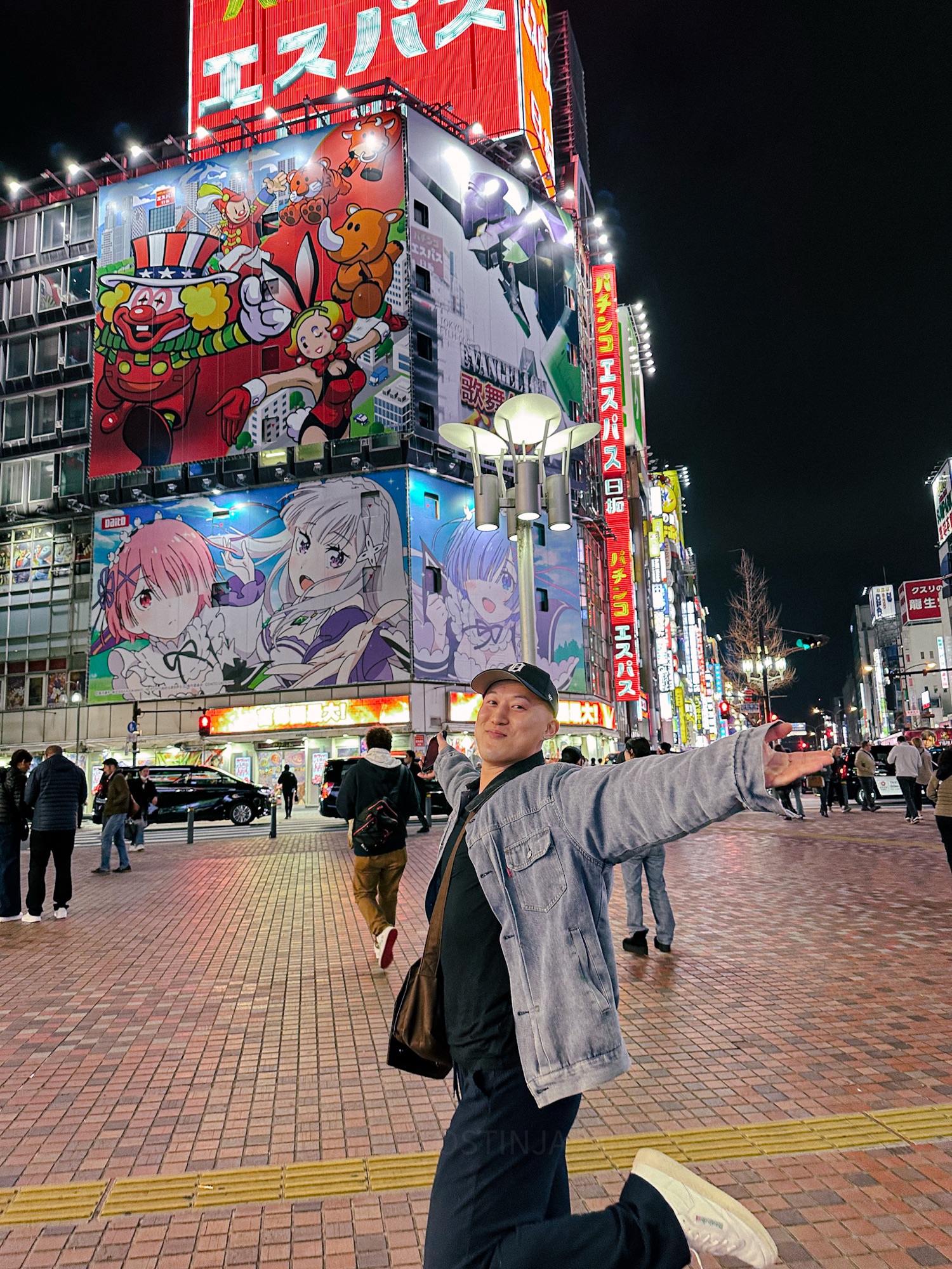 An Asian man standing with both arms raised up and a large smile on his face. One leg is kicked up as he danced in Akihabara, Tokyo's electric anime area. There is a large building with anime cartoon girls on the front.