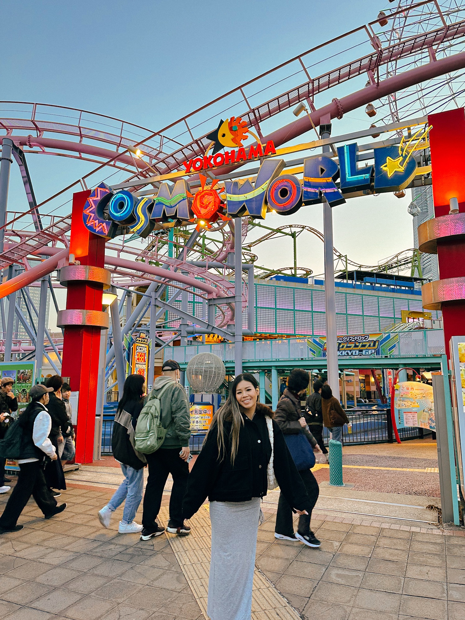 A girl in a black jacket and gray dress smiling under a sign that says "Cosmo World". Cosmo World is an attraction in Yokohama, the second largest city in Japan and an easy day trip from Tokyo.