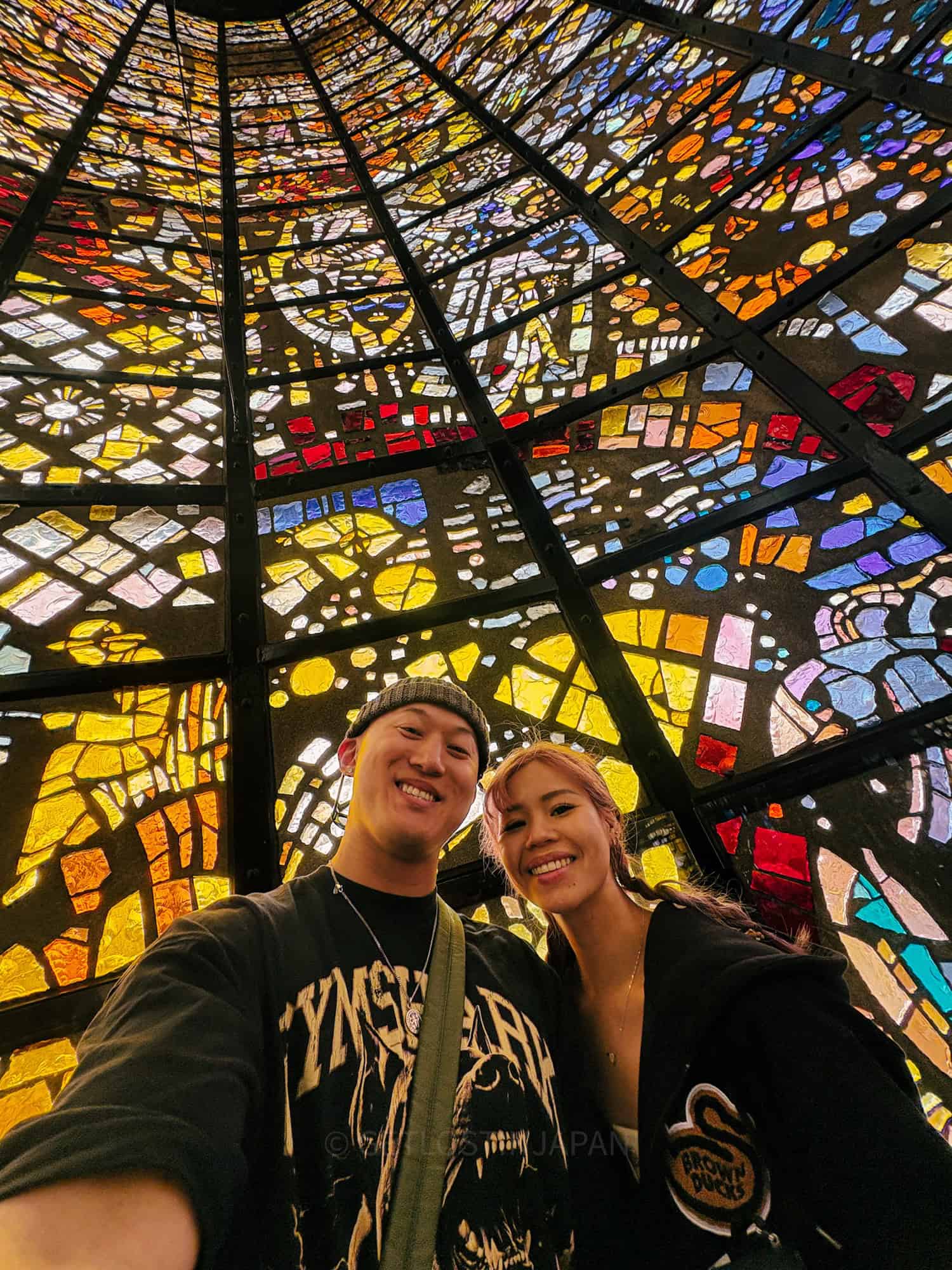 A man wearing a beanie and a woman with pink hair smiling in a stained glass building in the Hakone Open Air Museum.