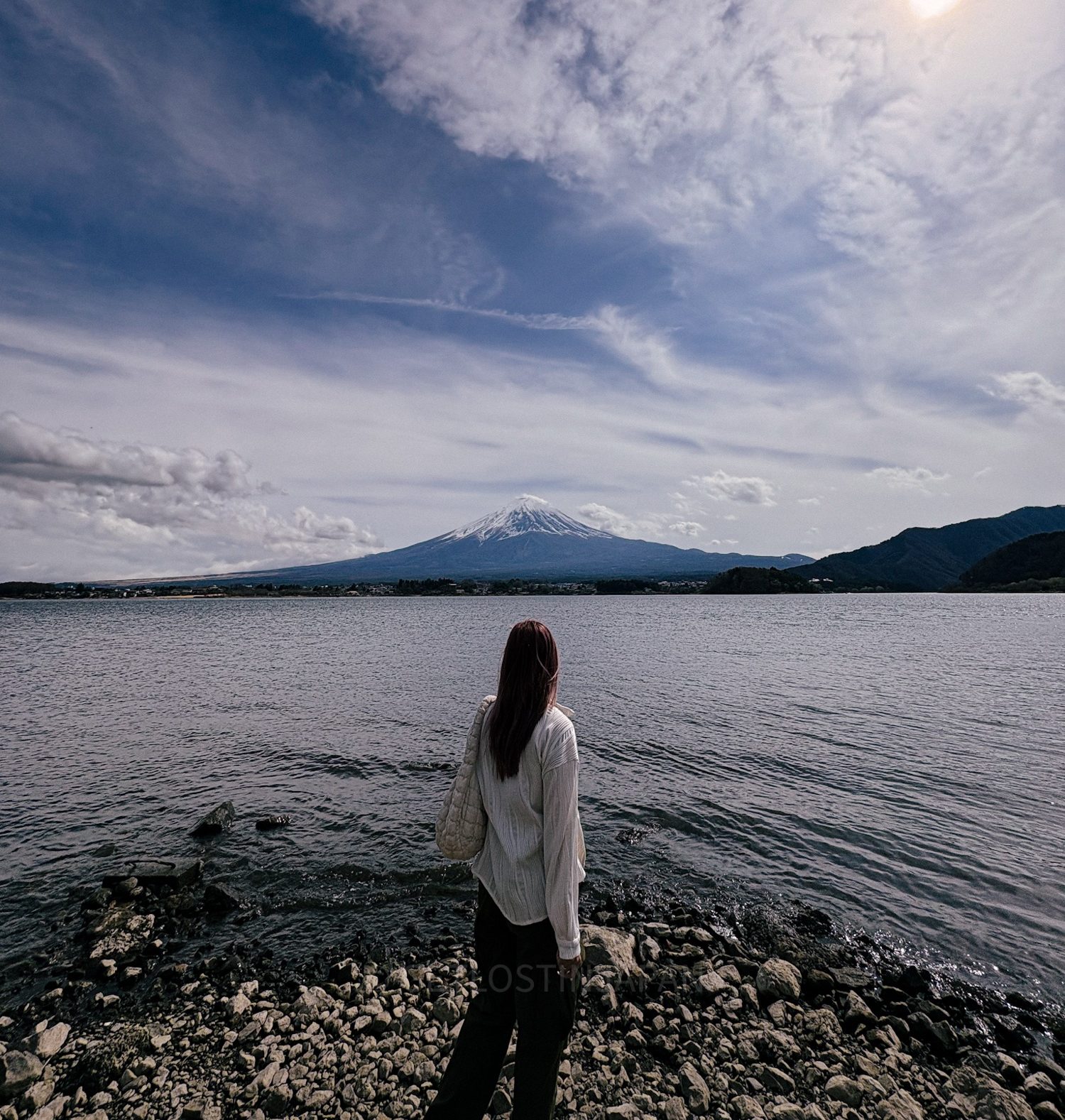 A girl in a white shirt and jeans looking at Mount Fuji across Lake Kawaguchi on a clear sunny day.