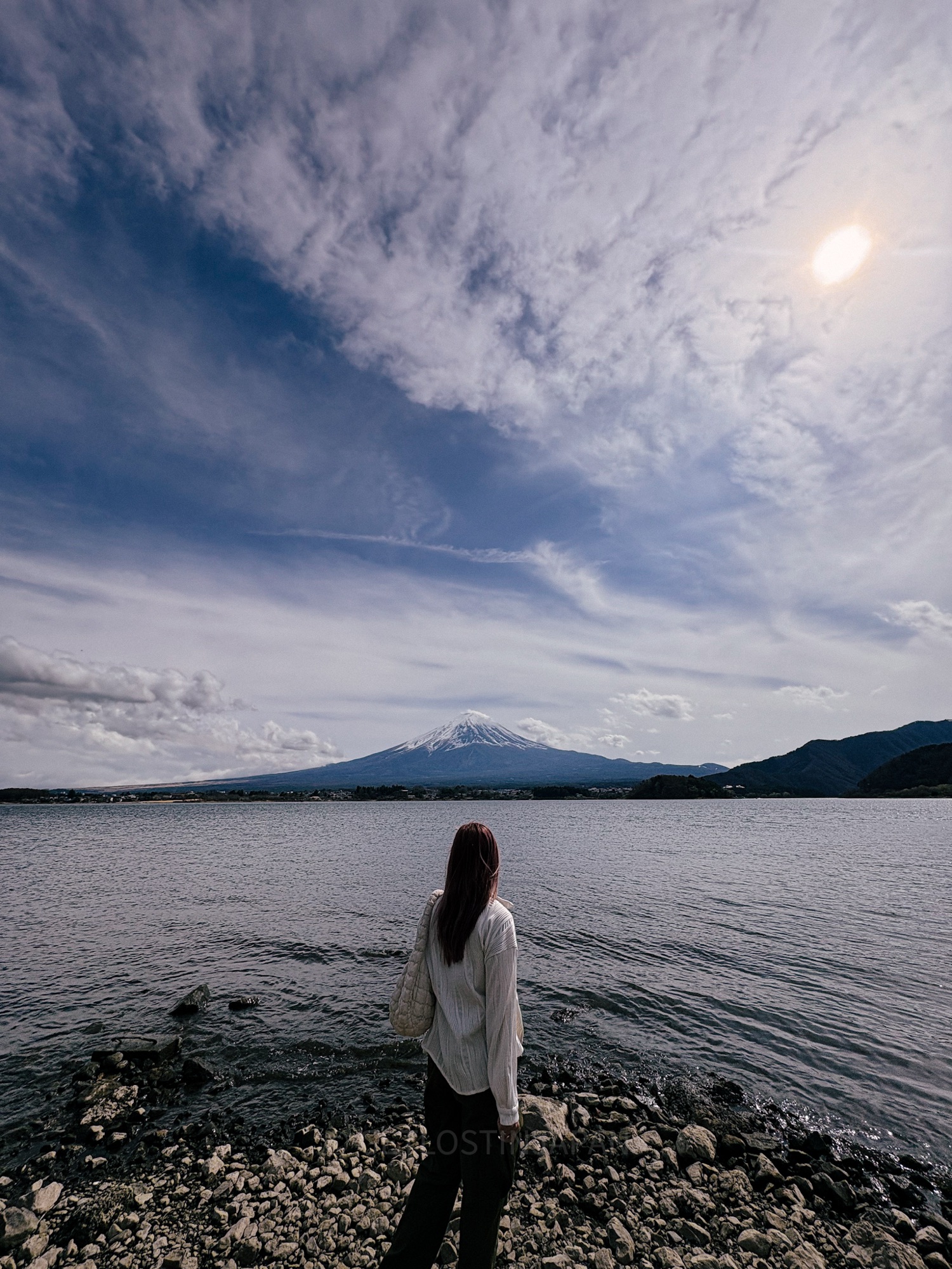 A girl in a white shirt and jeans looking at Mount Fuji across Lake Kawaguchi on a clear sunny day.