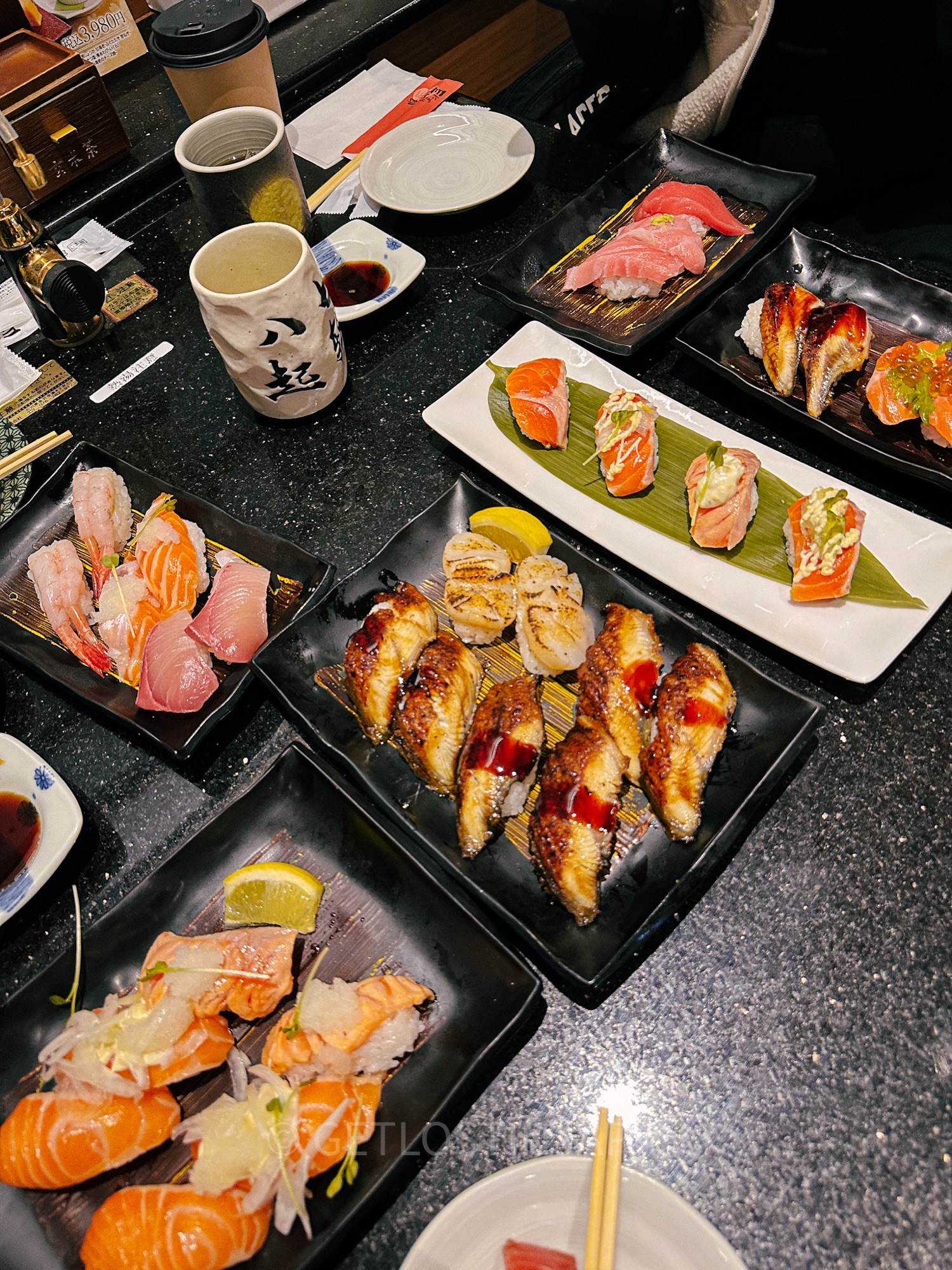 A table is filled with an assortment of sushi. Colorful fish such as tuna and salmon are topped with unagi sauce and spicy mayonnaise.