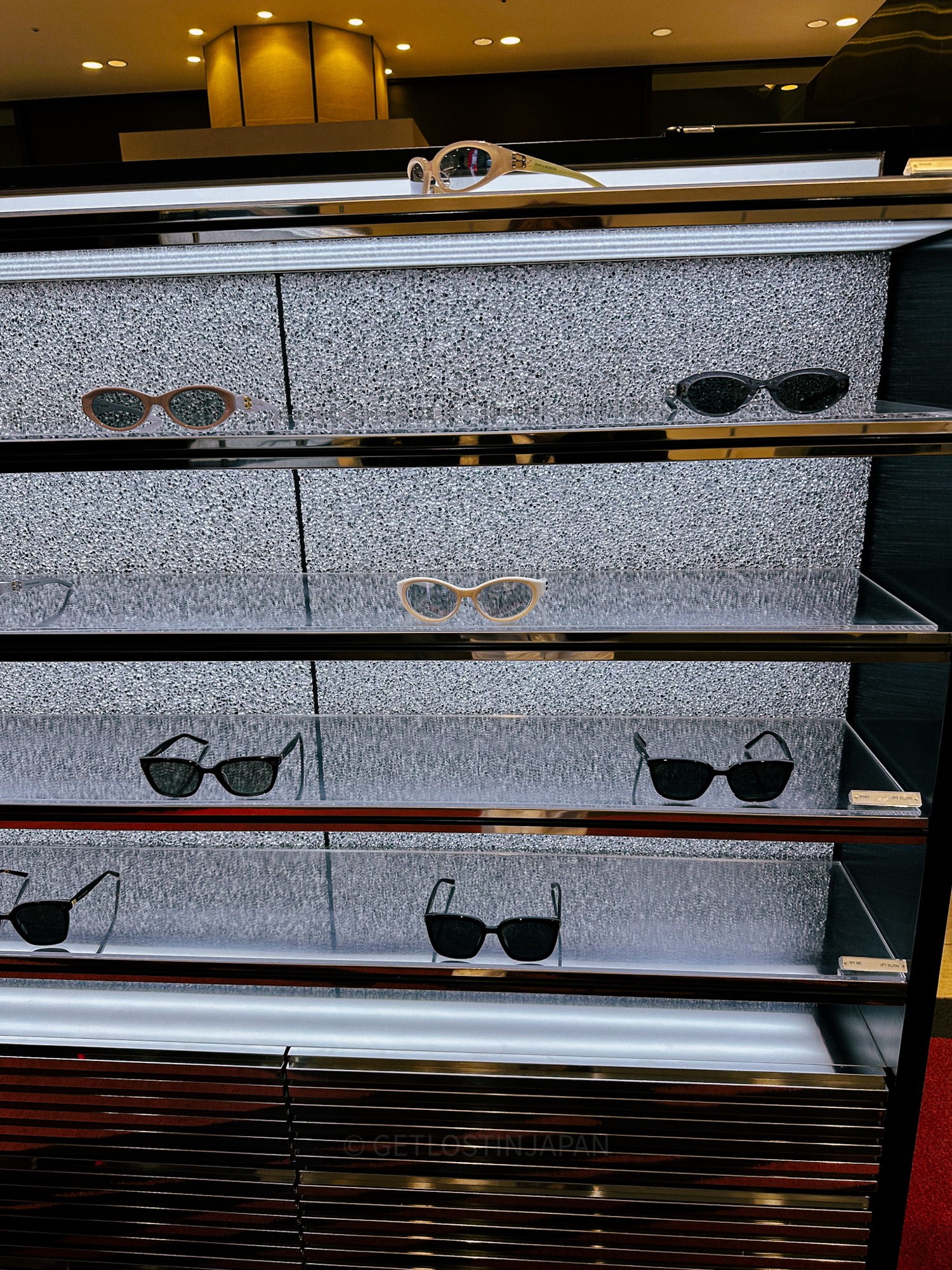A display of sunglasses at Gentle Monster in Hankyu Mall in Osaka. The glasses are illuminated with cool, white light and are displayed on a sleek modern grey shelf.