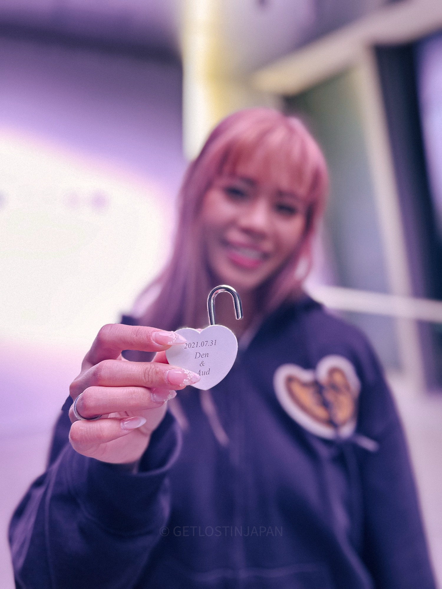 A girl with pink heart holds up a metal lock, exclusively purchased at Umeda Sky. The lock says 2021.07.31 Den & Aud
