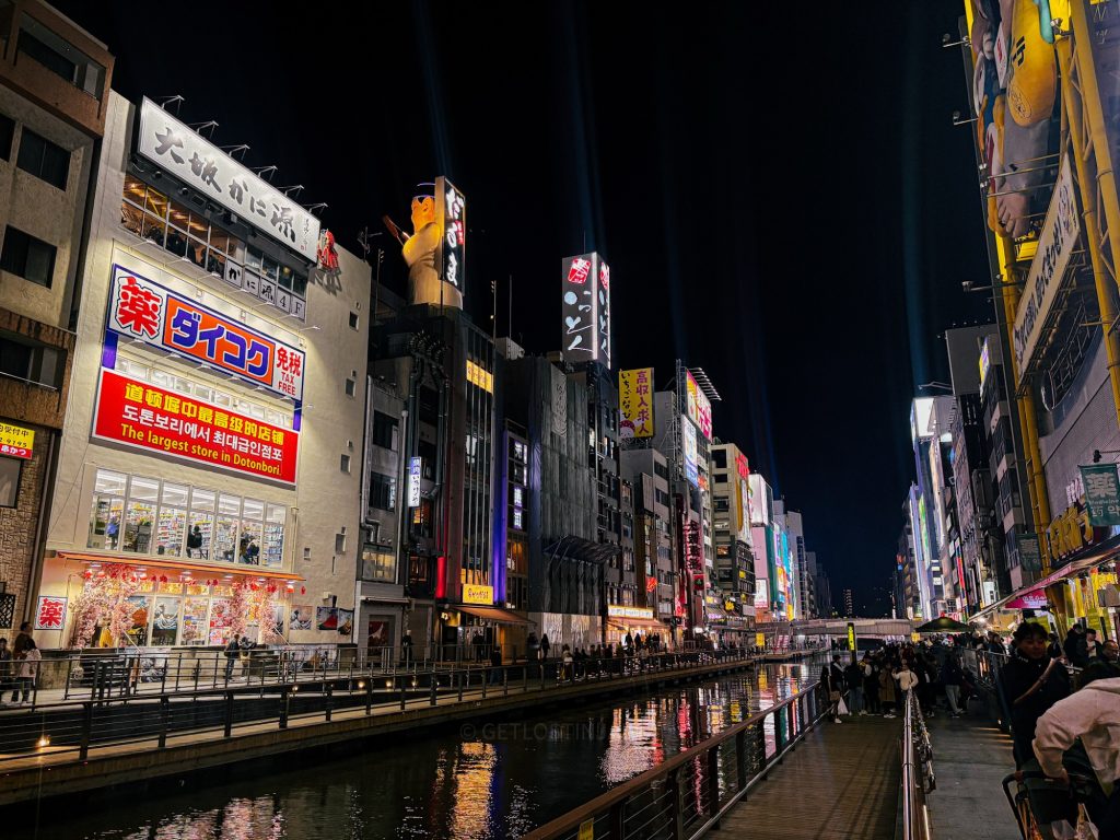 The Ebisu Bridge at night in Osaka, Japan. There are many shops and restaurants reflecting light on the calm water, and the street is bustling with people.