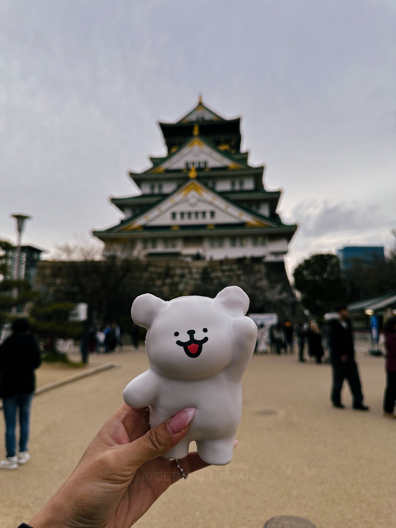 A white bear stuffed animal being held up in one hand, with Osaka Castle in the background during a cloudy day.