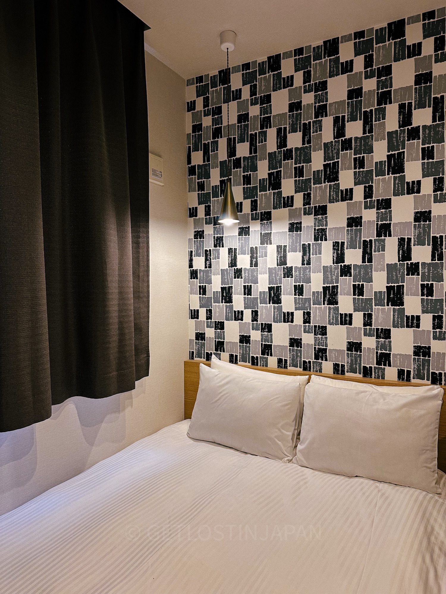 A hotel room in M's Hotel Gojo Odawara in Kyoto, Japan. There is a white double bed with blue, green, and white squared wallpaper behind it.
