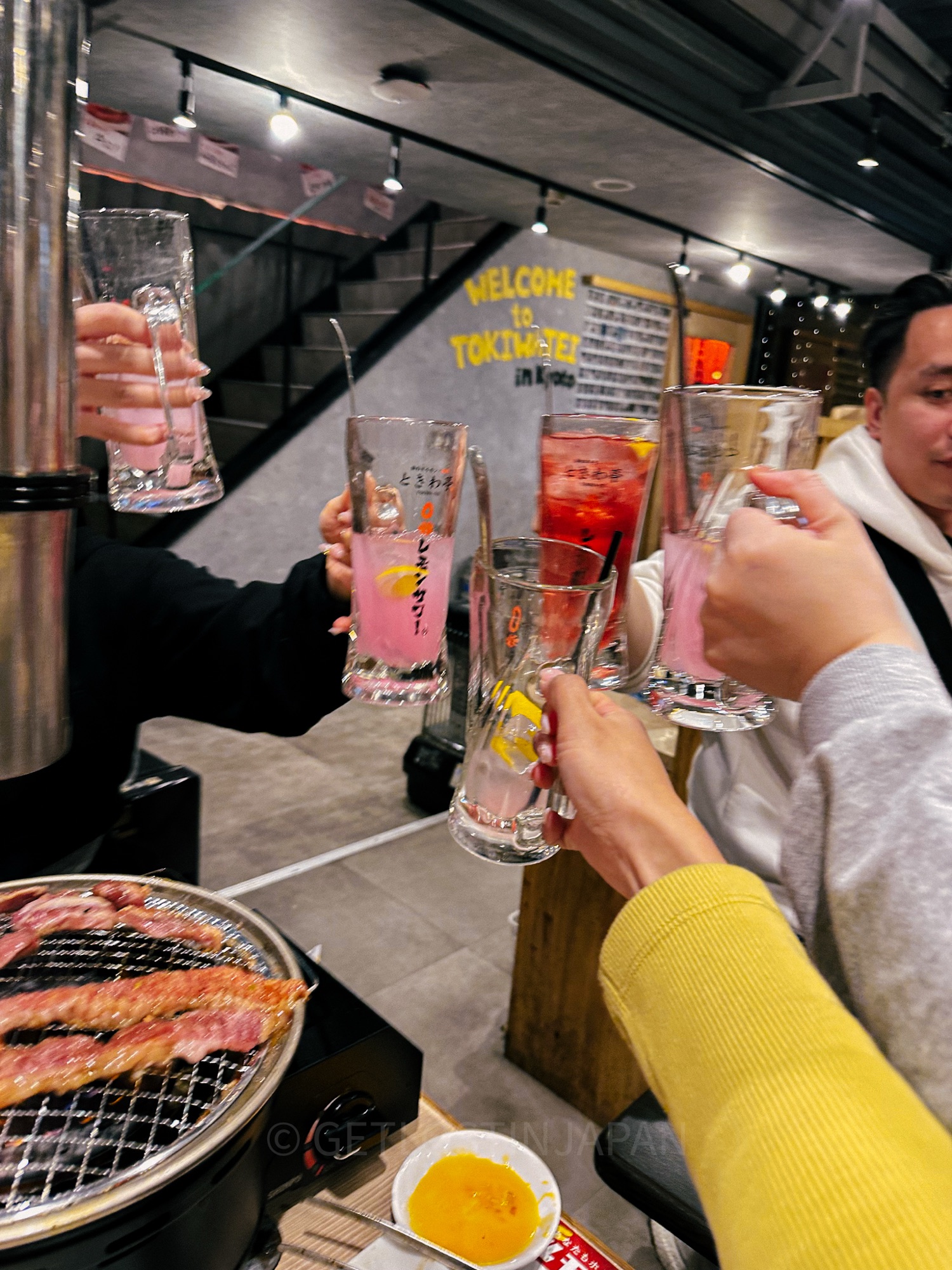 A group of people cheering in a yakiniku Japanese barbecue restaurant. There are 6 beer glasses, with meat grilling on a circular grill in the corner.
