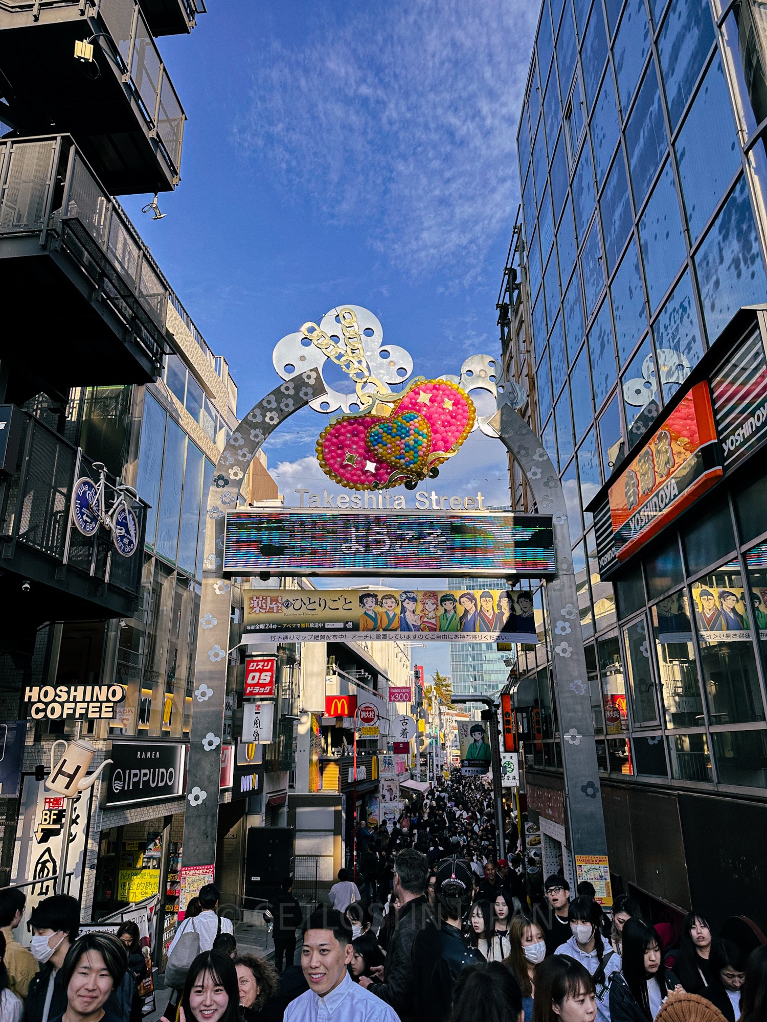 The entrance to Takeshita Street in Harajuku, Tokyo. This bustling street is filled with people, and a sign with colorful hearts reads the name of the street. Buildings surround the street on both sides on this clear sunny day.