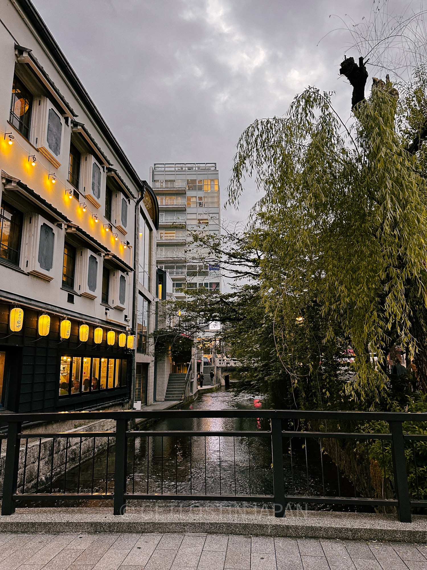 A bridge in Gion, the geisha district of Kyoto, Japan. A building with brightly shining yellow lanterns stands to the left of the bridge, while trees grow across the water to the right.