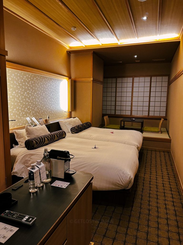 A hotel room in Saka Hotel Kyoto, a modern hotel that incorporates Japanese features such as wooden screen windows and earthy colors to create a calm environment. Two double beds are prepared with fresh white bedding, and free tea and coffee sit on the table.