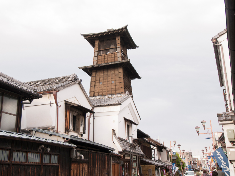 Kawagoe, known as little Edo for its preserved buildings from the Edo period. The Time of Bell Tower, a large brown clock tower rests in the background of the picture, with bustling buildings all around.