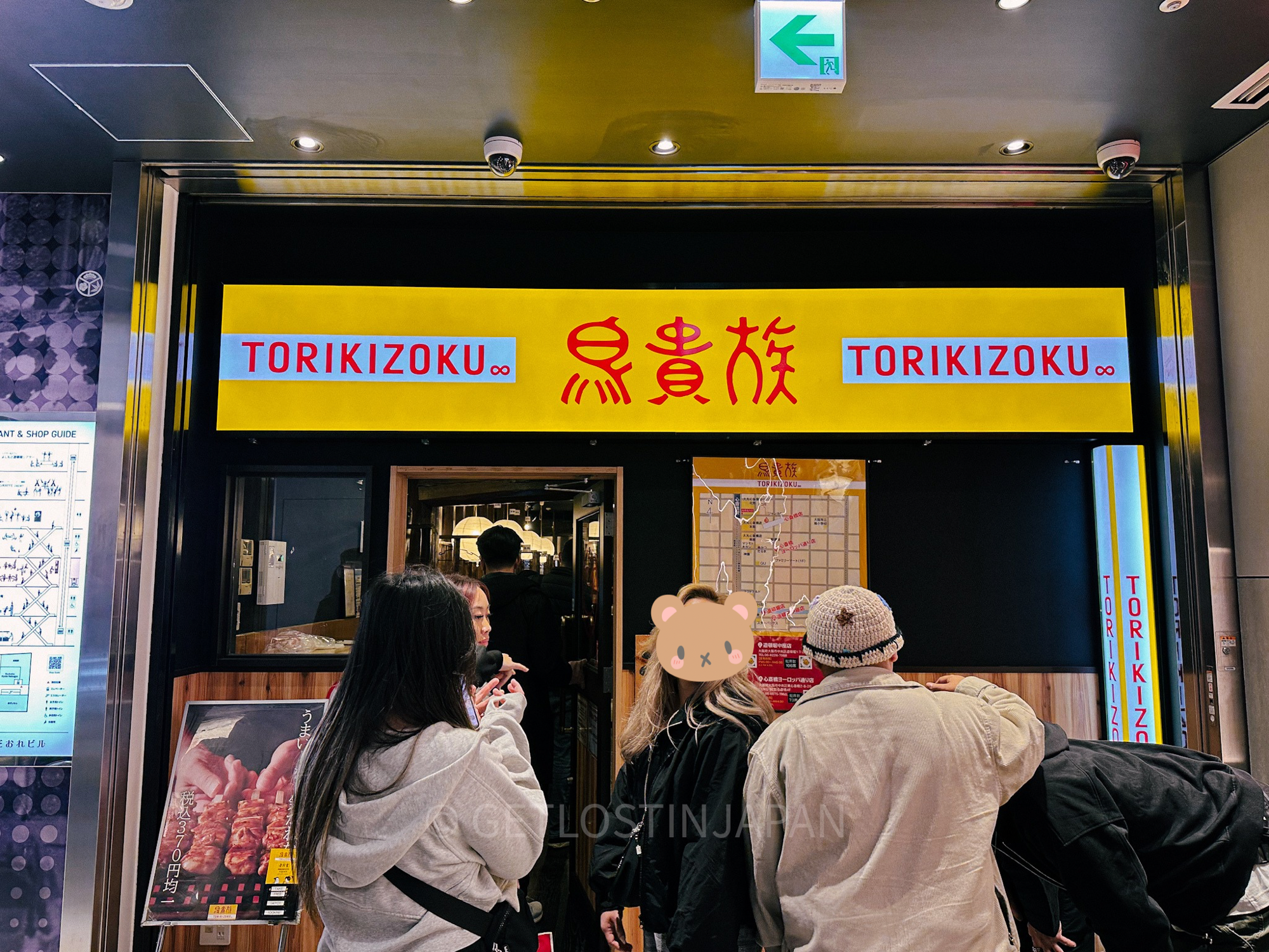 Three people enter a yakitori restaurant serving Japanese skewered foods. There is a large yellow sign with the words "Torikizoku" on each side.