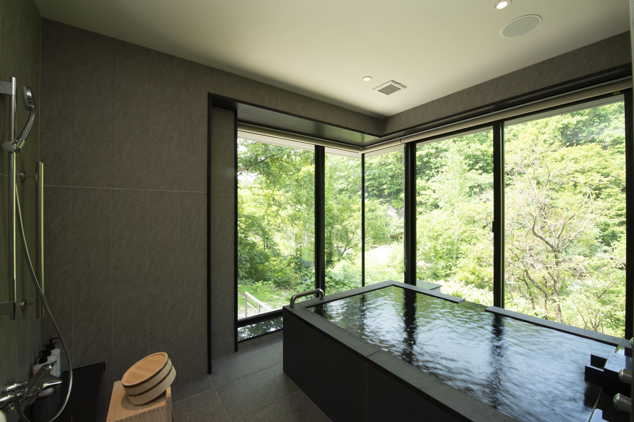 Private indoor onsen bath at Chalet Ivy Jozankei with sleek stone tiles and tranquil views of lush green trees through wraparound glass walls.