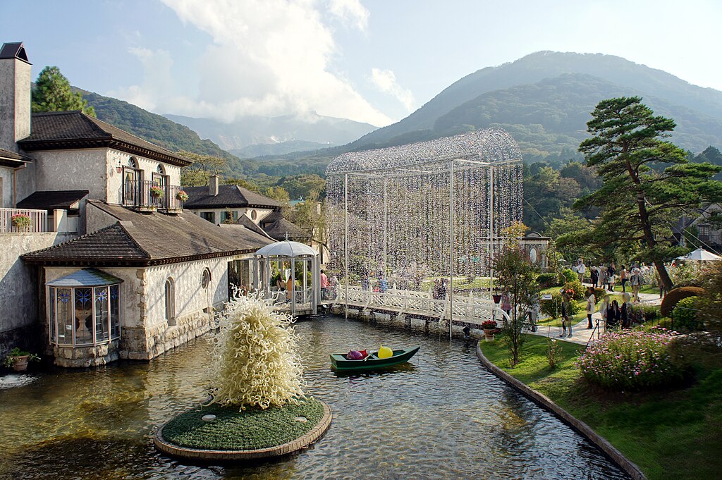 The Venetian Glass Museum in Hakone featuring European-style architecture, a sparkling crystal bridge over a pond, and Mount Hakone in the background on a sunny day.
