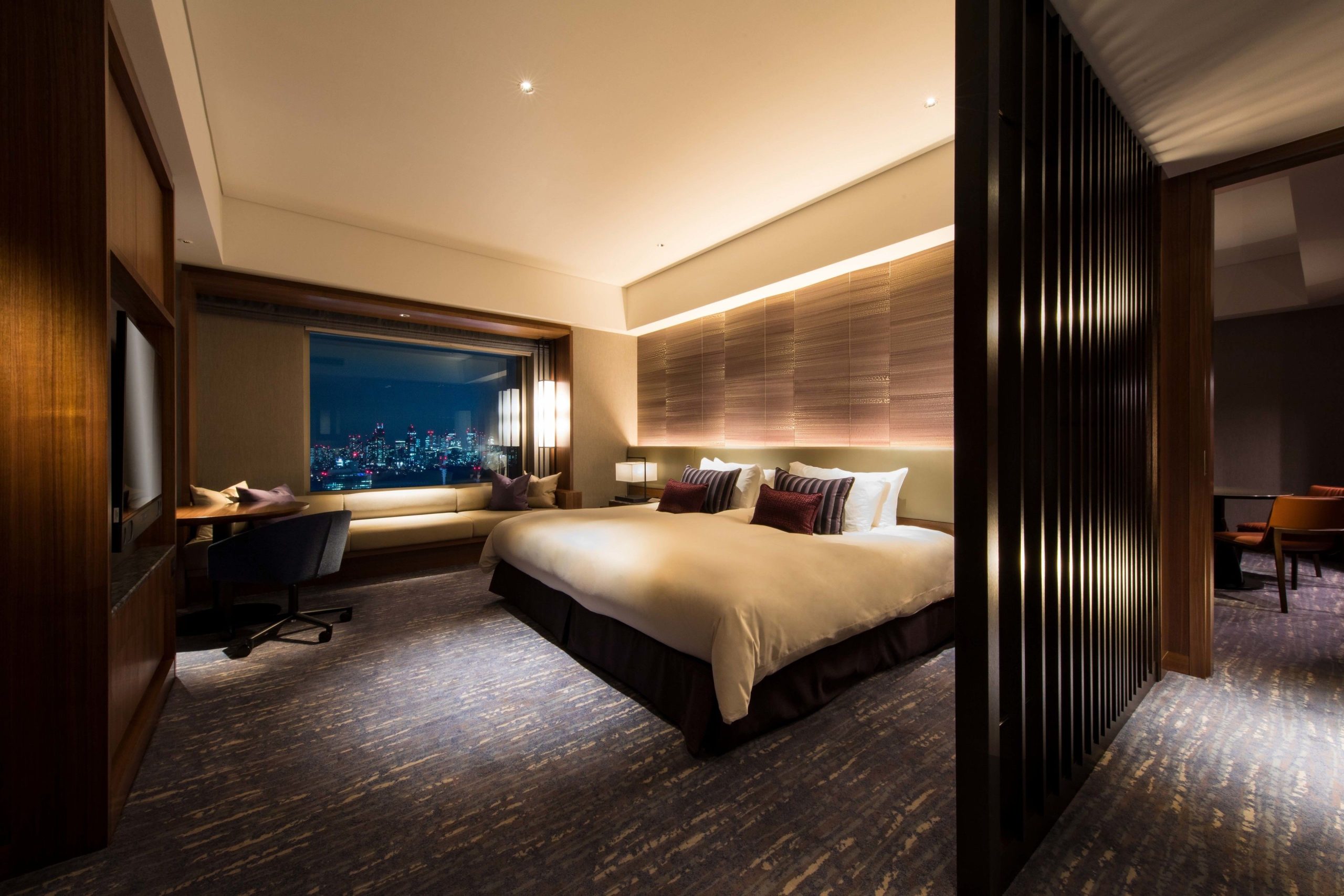 Elegant hotel room in Cerulean Tower with a large bed, cozy lighting, and floor-to-ceiling windows overlooking Shibuya's night skyline. A long sofa and modern desk sit by the window, creating a warm, luxurious atmosphere.