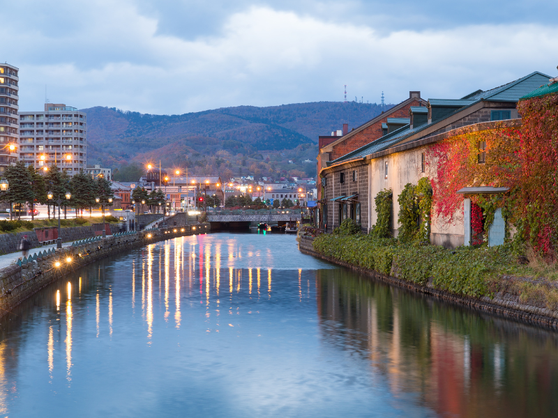 Evening view of the Otaru Canal with historic brick warehouses reflected in the water and soft lantern lights glowing — a must-see stop on an Otaru day trip.