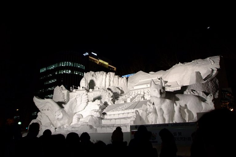 A large illuminated snow sculpture at the Sapporo Snow Festival featuring animals and buildings carved in detailed relief, with visitors standing in front at night.